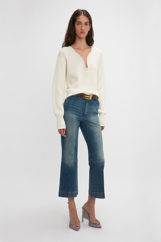 A woman stands in a white studio, wearing a cream knit sweater, belted Victoria Beckham cropped kick jeans in Indigrey wash, and gray high-heeled sandals.