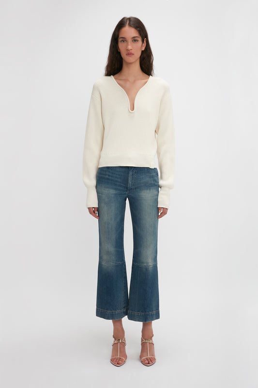 A woman stands against a plain background, wearing a white V-neck sweater, blue cropped jeans, and Victoria Beckham Frame Detail Sandal In Nude Leather. She has long, loose hair.