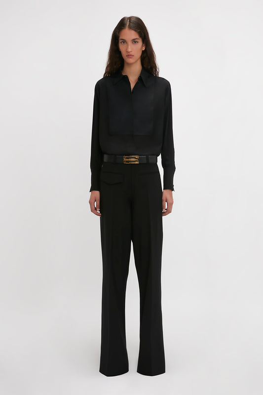 A person stands facing forward, showcasing a sleek contemporary silhouette. They're dressed in a black button-up shirt and Victoria Beckham Reverse Front Trouser In Black with a gold-buckled belt. The background is plain white.
