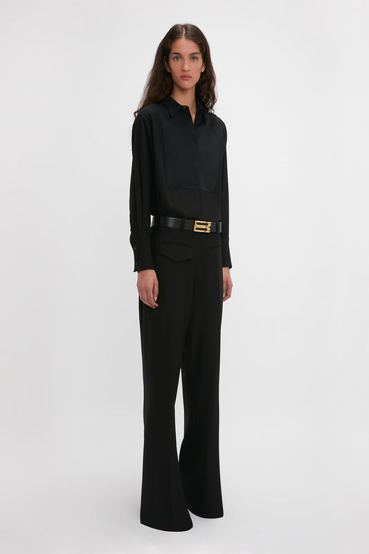 Woman in black outfit: loose-fitting blouse tucked into Victoria Beckham's Reverse Front Trouser In Black, accessorized with a wide belt. She embodies the modern woman, standing against a plain white background with a contemporary silhouette.