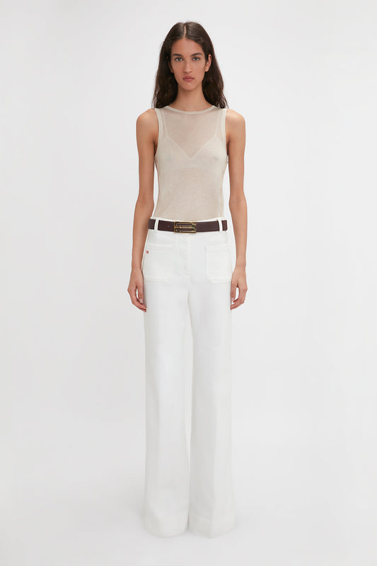 A person with long hair is standing against a plain backdrop, wearing a Lightweight Tank Top In Birch by Victoria Beckham and high-waisted white pants with a brown belt.