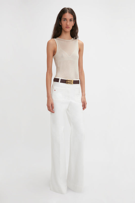 A woman with long dark hair wears a sleeveless, sheer beige top and high-waisted white wide-leg pants; she has a black belt with a gold buckle. She stands against a plain white background. She is wearing the Lightweight Tank Top In Birch by Victoria Beckham.