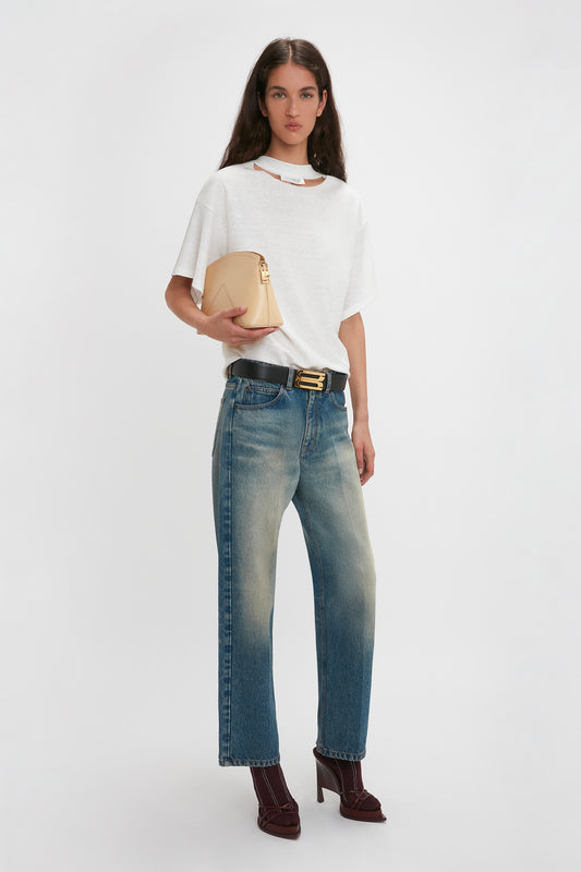 A woman wearing a white T-shirt, faded blue jeans, and burgundy high-heeled shoes stands holding a versatile Victoria Beckham Victoria Clutch Bag In Sesame Leather.