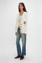 Person standing against a plain background, wearing a Relaxed Fit Cardigan In Natural by Victoria Beckham over a white top, blue jeans, and brown shoes.