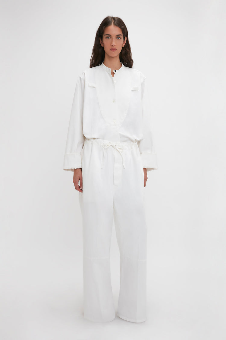 A woman in a white cotton-canvas shirt and Victoria Beckham's Drawstring Pyjama Trousers In Washed White stands against a white background, looking directly at the camera.