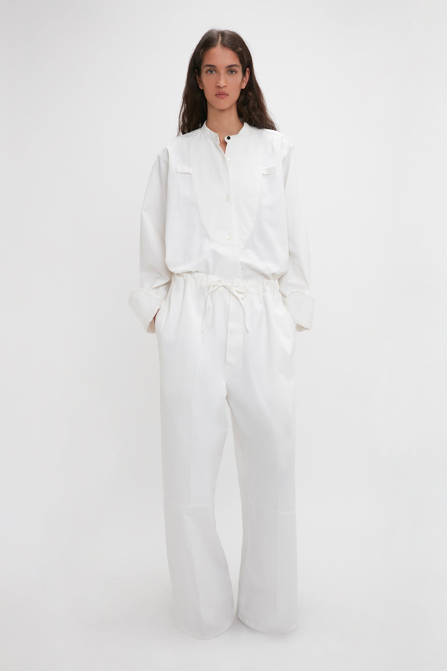 A woman in Victoria Beckham's drawstring pyjama trouser in washed white, with wide-leg trousers and cinched waist, standing against a plain light background.