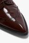 Close-up of the toe of a polished, dark brown, calfskin leather Victoria Beckham Flat Lace Up Mules In Bordeaux Leather with decorative perforations and stitching detail.