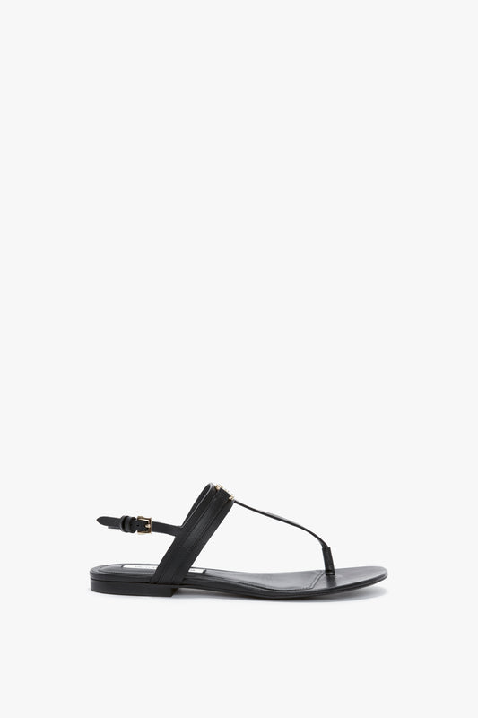 Black flat sandal with a strappy design, featuring a thong-style toe strap and adjustable ankle buckle on a white background. The Victoria Beckham Flat Chain Sandal In Black Leather is crafted from premium calfskin leather, offering both style and comfort.
