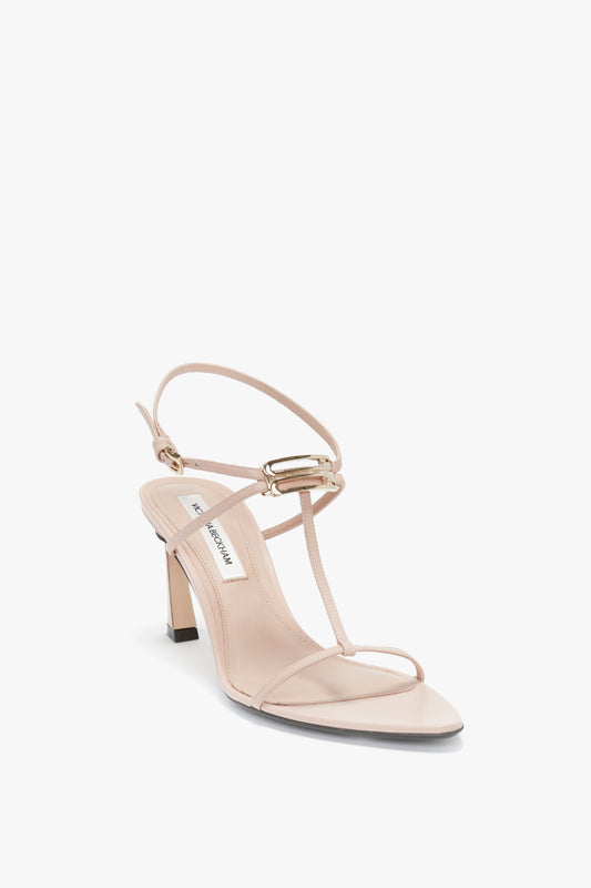 A single Victoria Beckham Frame Detail Sandal In Nude Leather with a thin, adjustable ankle strap, front strap, and small gold buckle. It features a V-shaped sculptural heel and is displayed against a white background.
