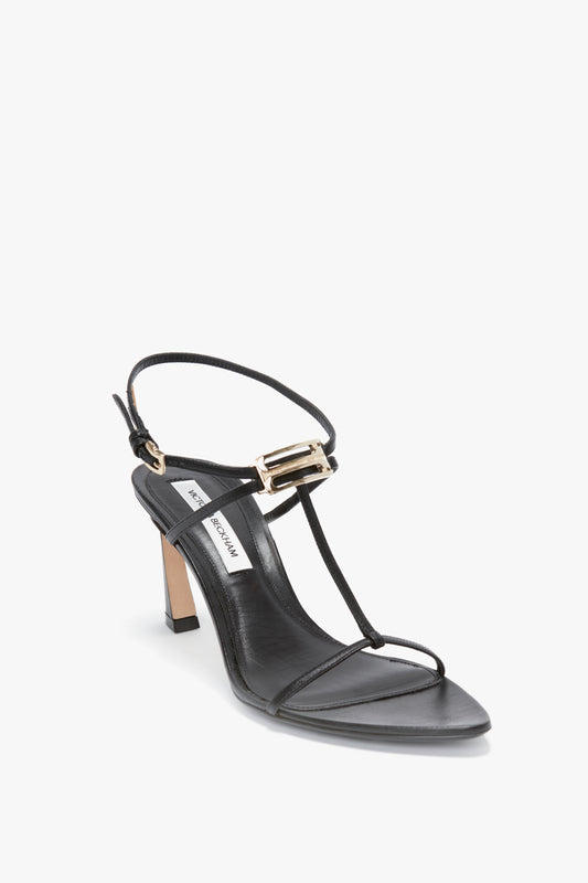 The Frame Detail Sandal In Black Leather by Victoria Beckham features thin straps, a T-strap design, an adjustable ankle strap with a gold buckle, and a sculptural heel. The insole is labeled "Victoria Beckham.