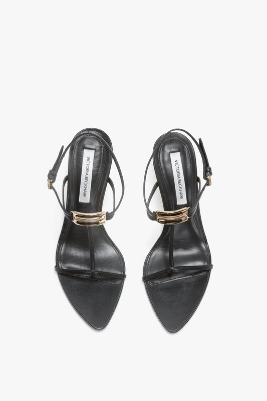 A pair of Victoria Beckham Frame Detail Sandal In Black Leather with thin straps and metallic buckle accents, featuring an adjustable ankle strap and sculptural heel, all on a white background.