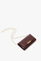 A maroon leather handbag with a gold chain strap and a gold clasp closure, lying against a white background, reminiscent of the elegance of the Wallet On Chain In Burgundy Leather by Victoria Beckham.