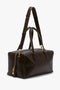 A sleek dark brown leather duffel bag with gold hardware, adjustable shoulder strap, and double handles, the Medium Gym Bag In Chocolate Brown by Victoria Beckham features an exterior zip pocket on one side.