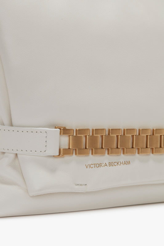 Close-up of a white Puffy Chain Pouch With Strap In White Leather with a prominent gold chain detail and the brand's name embossed in gold lettering on the bag's surface, reminiscent of Victoria Beckham’s vintage watch straps.