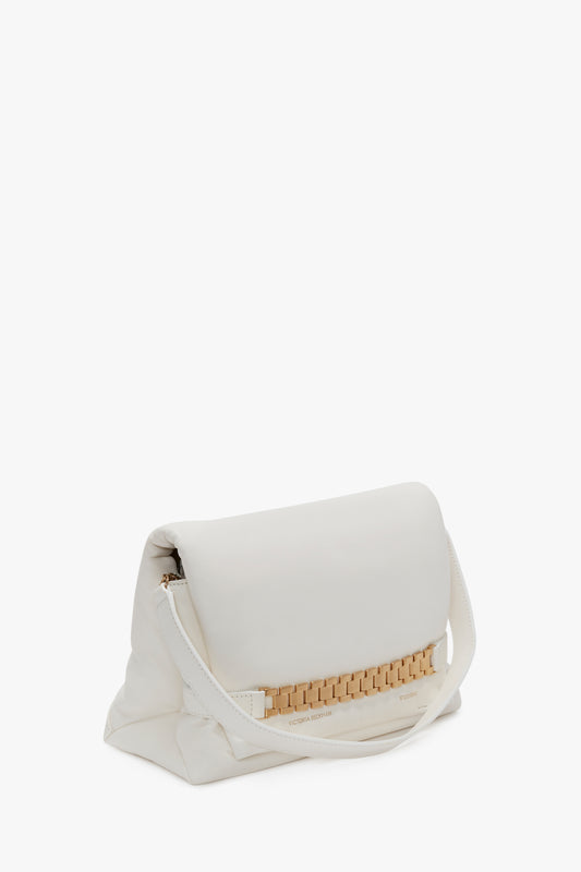 A white, folded Puffy Chain Pouch With Strap In White Leather by Victoria Beckham has a cream strap and decorative gold detail at the base. The bag has a minimalistic design, smooth texture, and evokes the elegance of Victoria’s vintage watch straps.
