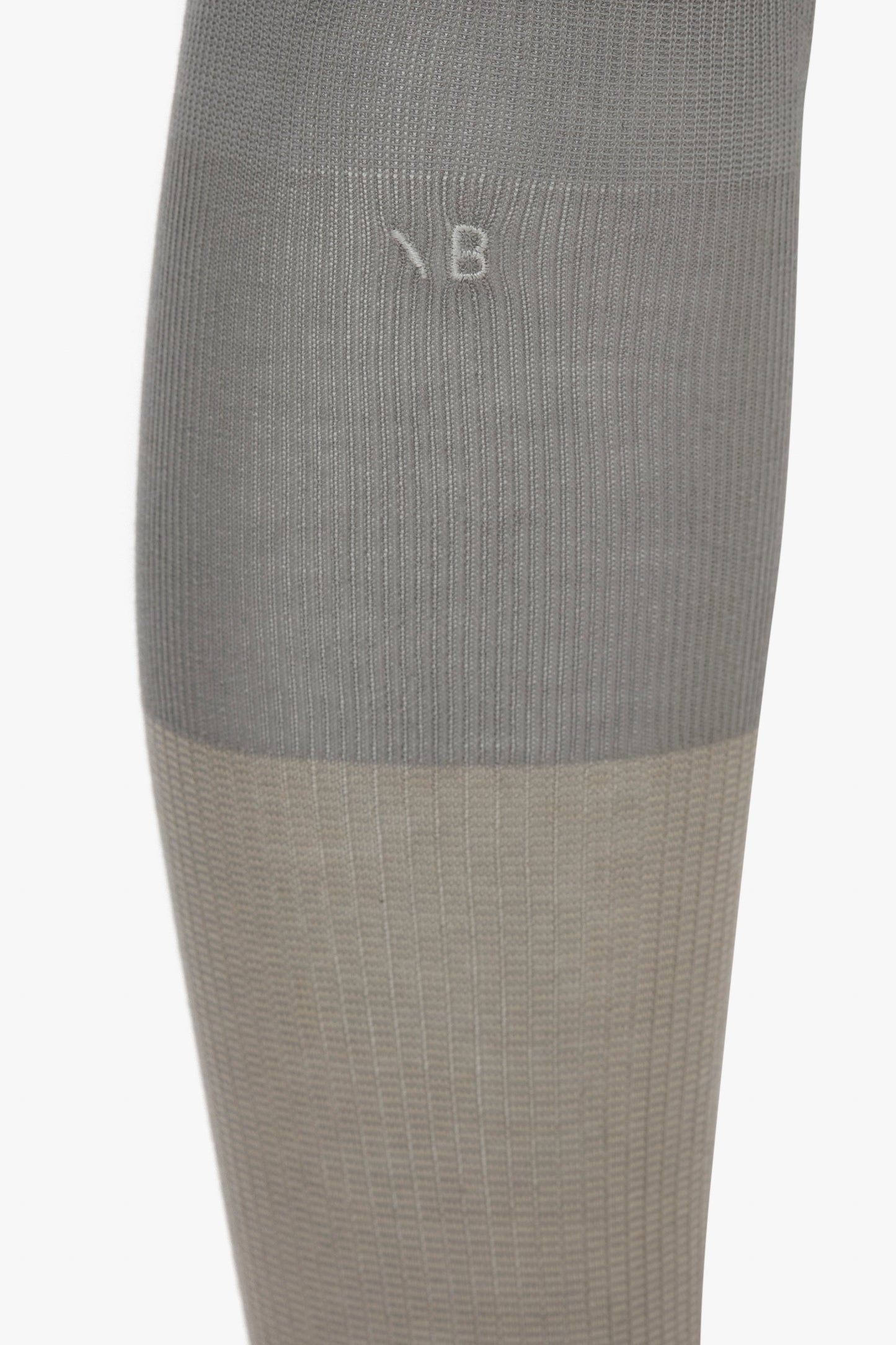 Close-up of a gray, ribbed-knit garment made from 100% cotton with a contrasting Lunar Grey and Sesame section and a small white letter "B" stitched near the top of the image. The garment is part of Victoria Beckham's collection, specifically the Superfine Rib Socks In Lunar Grey.