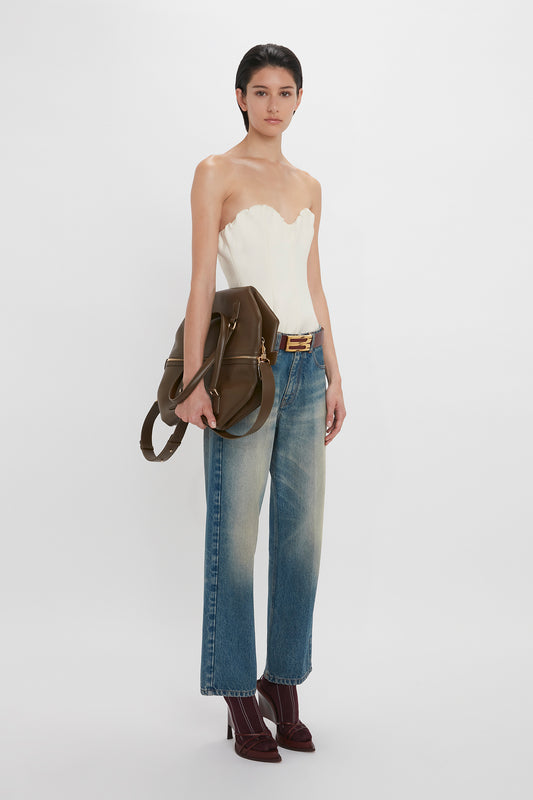 A woman stands against a white backdrop, wearing a Victoria Beckham corset top in antique white with a sweetheart neckline, blue jeans with a belt, and brown boots, holding a brown shoulder bag.