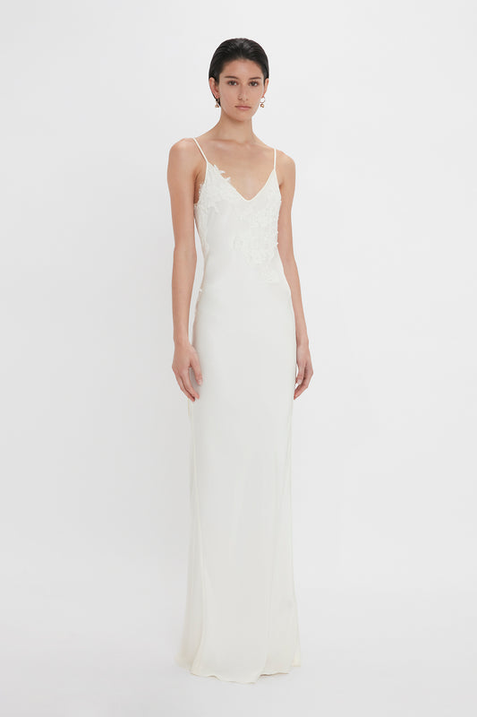 A woman in a sleek, white sleeveless bridal gown with lace appliqué on the bodice, standing against a plain white background wearing the Victoria Beckham Exclusive Lace Detail Floor-Length Cami Dress In Ivory.