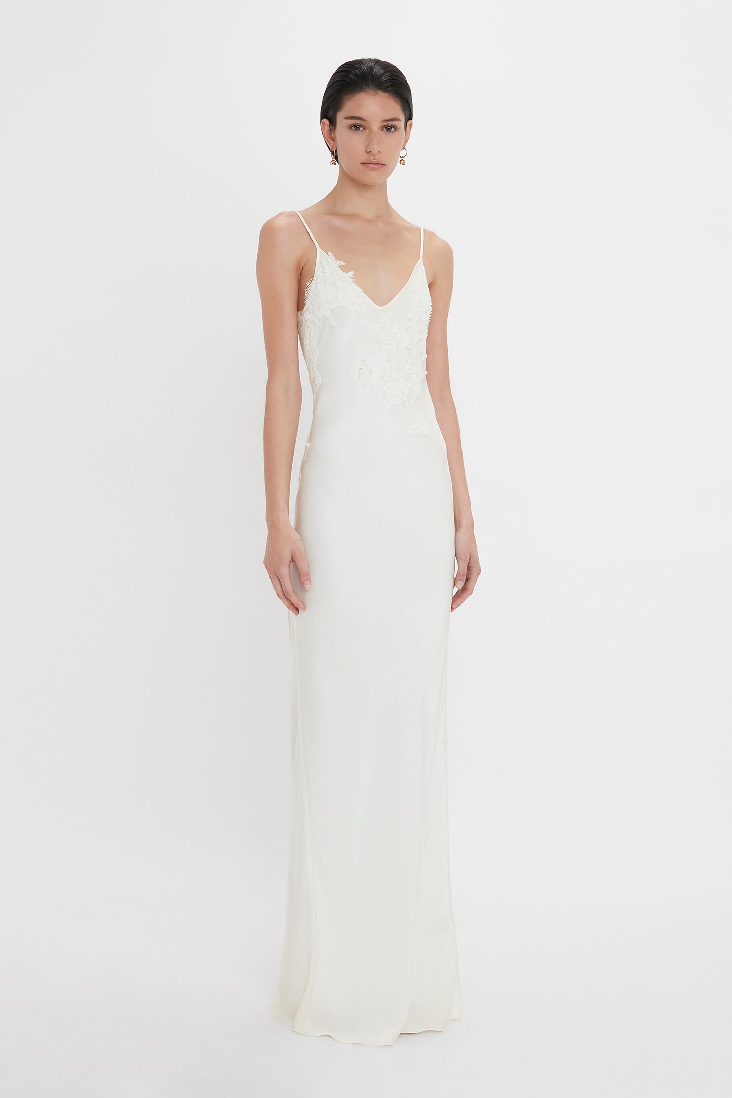 A woman in a sleek, white sleeveless wedding dress with lace detailing at the bodice, standing against a plain white background, wearing Victoria Beckham's Exclusive Camellia Flower Hoop Earrings In Gold.