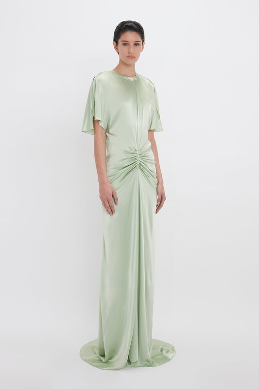 A woman in a Victoria Beckham Exclusive Floor-Length Gathered Dress In Jade, with an open back and a twisted detail at the waist, standing against a plain white background.