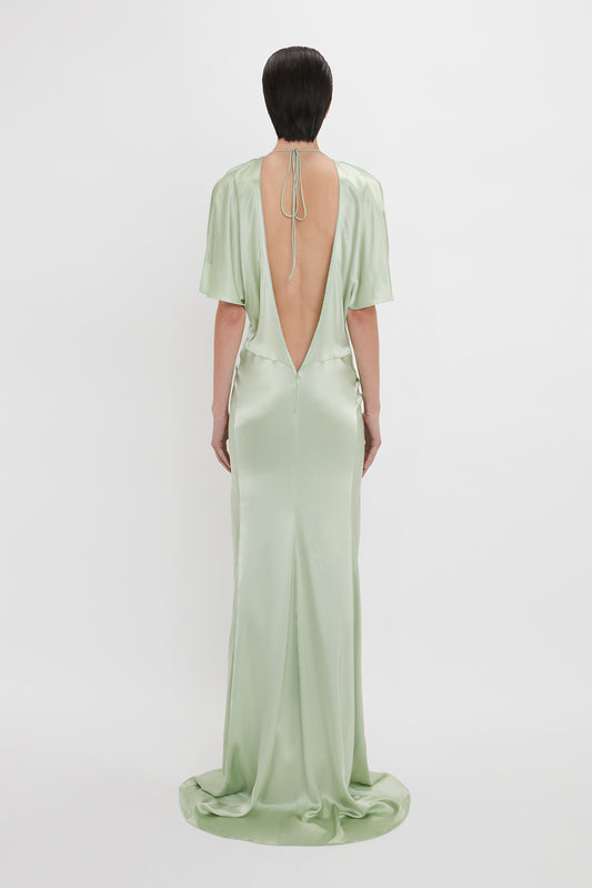 A woman stands facing away, wearing an elegant Victoria Beckham Exclusive Floor-Length Gathered Dress in Jade with an open back and a small train, her hair styled in a sleek bun.