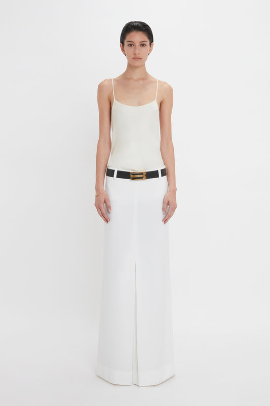 A person stands against a plain white background wearing an Exclusive Cami Top In Ivory by Victoria Beckham, a white floor-length skirt, and a black belt.