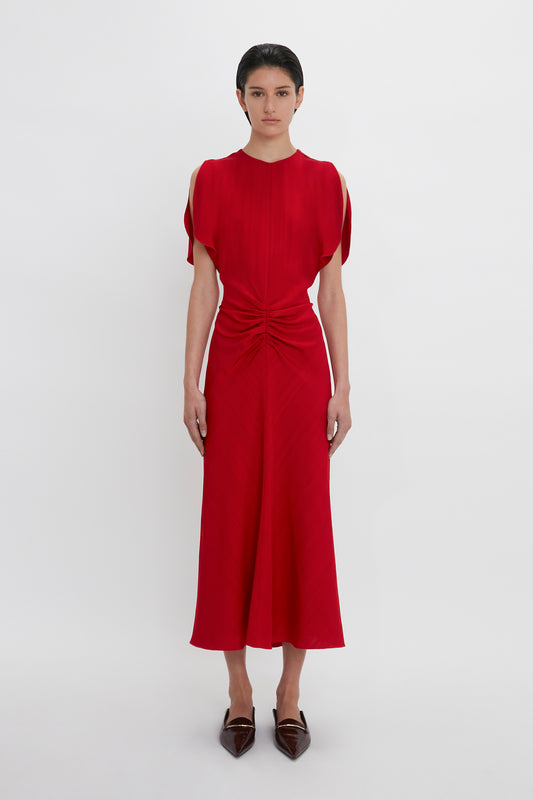 A woman in a red Exclusive Gathered V-Neck Midi Dress In Carmine with cap sleeves and a knotted detail at the waist stands against a plain white background, wearing dark loafers by Victoria Beckham.