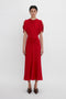 A woman in a red Exclusive Gathered V-Neck Midi Dress In Carmine with cap sleeves and a knotted detail at the waist stands against a plain white background, wearing dark loafers by Victoria Beckham.