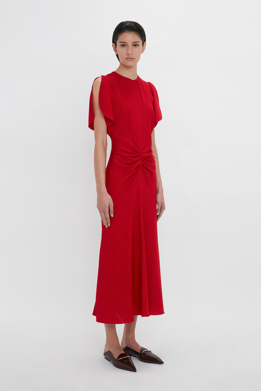 A woman in an Exclusive Gathered V-Neck Midi Dress In Carmine by Victoria Beckham, standing against a plain white background, wearing dark loafers.