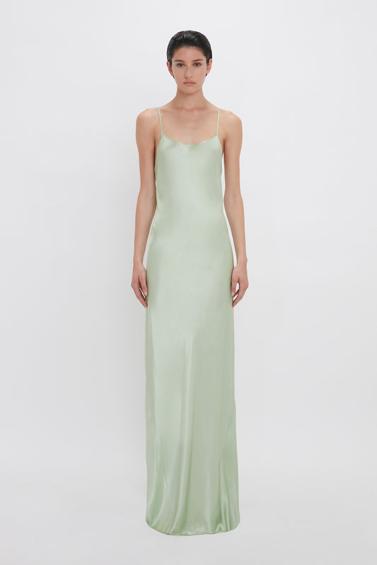 A woman in a flowing, light green, crepe back satin Exclusive Low Back Cami Floor-Length Dress In Jade standing against a plain white background by Victoria Beckham.