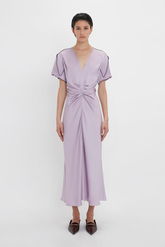 Person wearing the Gathered V-Neck Midi Dress In Petunia by Victoria Beckham with short sleeves and a waist-defining pleat, paired with pointed brown shoes. They are standing against a plain white background.