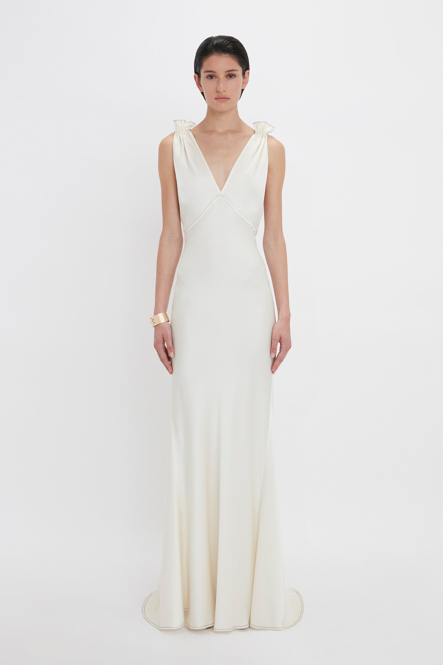 A person stands facing forward, wearing a sleeveless, floor-length Gathered Shoulder Floor-Length Cami Gown In Ivory by Victoria Beckham made of crepe back satin with a plunging V-neckline. They have short, dark hair and are accessorized with a bracelet. The elegant ensemble captures the timeless allure of sophisticated fashion.