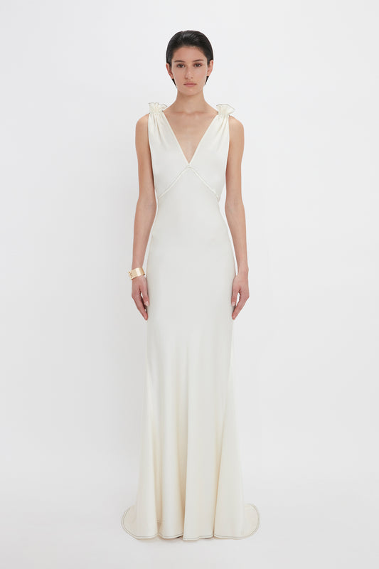A person stands facing forward, wearing a sleeveless, floor-length Gathered Shoulder Floor-Length Cami Gown In Ivory by Victoria Beckham made of crepe back satin with a plunging V-neckline. They have short, dark hair and are accessorized with a bracelet. The elegant ensemble captures the timeless allure of sophisticated fashion.