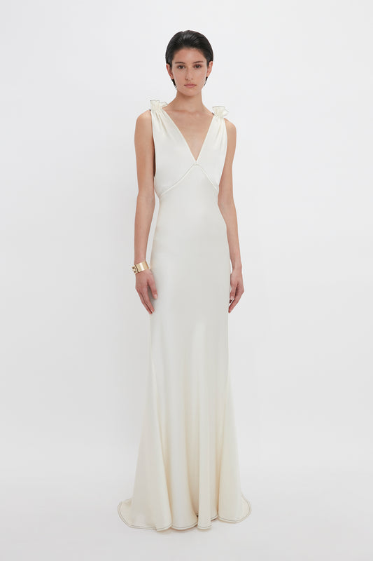 A person in a sleeveless, floor-length, backless Gathered Shoulder Floor-Length Cami Gown In Ivory by Victoria Beckham stands against a plain white background, wearing a gold bracelet.