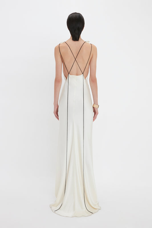 A person with short dark hair is wearing a Gathered Shoulder Floor-Length Cami Gown In Ivory by Victoria Beckham with thin black straps and standing against a white background, facing away from the camera.