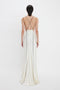 A person with short dark hair is wearing a Gathered Shoulder Floor-Length Cami Gown In Ivory by Victoria Beckham with thin black straps and standing against a white background, facing away from the camera.