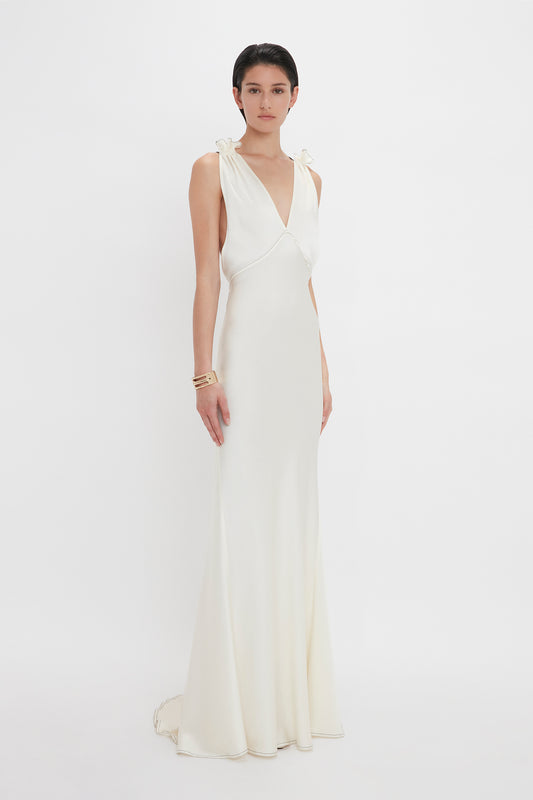 A person in a sleeveless, Victoria Beckham Gathered Shoulder Floor-Length Cami Gown In Ivory with a deep V-neckline stands against a plain white background. They are wearing bracelets on their right wrist.