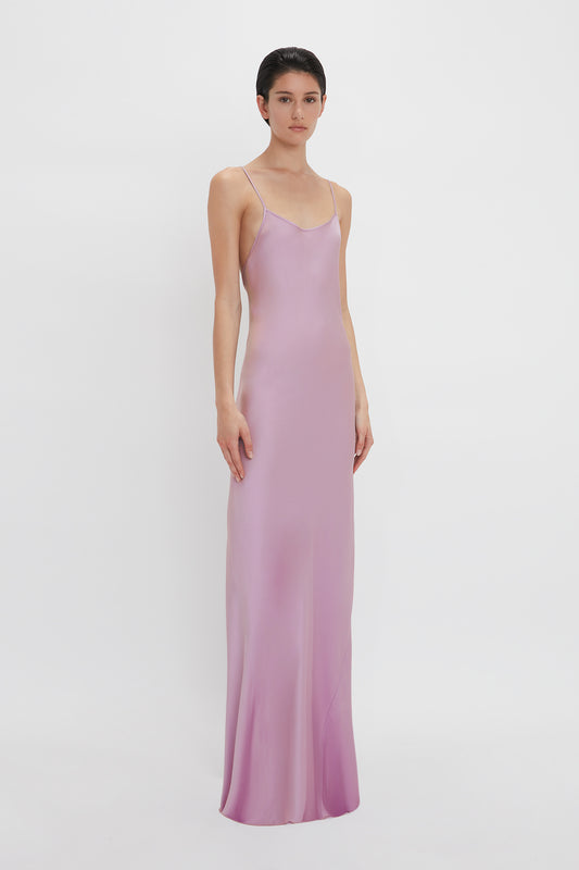 Woman standing against a white background wearing a long, elegant, light purple satin Low Back Cami Floor-Length Dress In Rosa slip dress with spaghetti straps by Victoria Beckham.