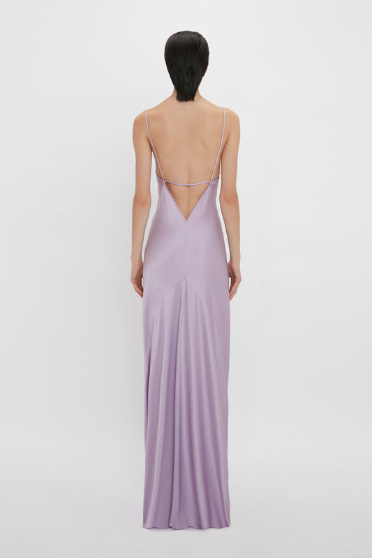 A woman viewed from behind, wearing a floor-length Low Back Cami Dress in Petunia by Victoria Beckham, with thin straps, standing against a white background.