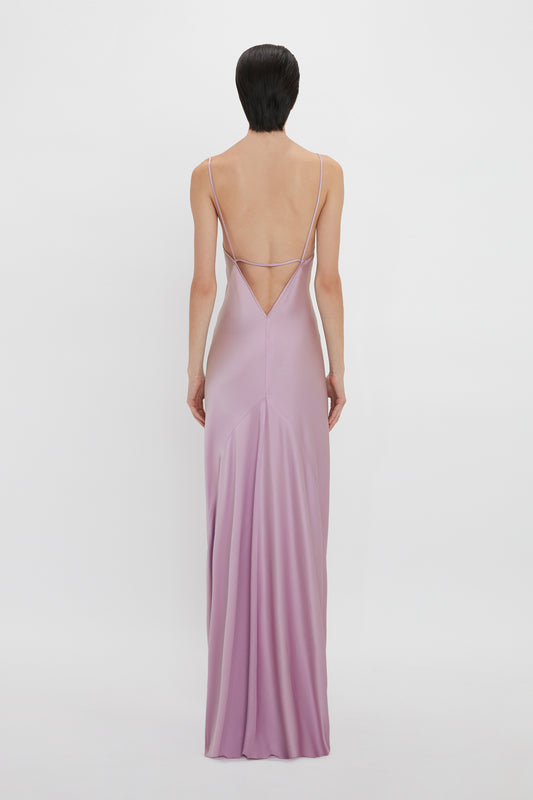 Rear view of a woman standing against a white background, wearing a long, elegant pastel purple camisole slip dress with thin straps and a crisscross back design from Victoria Beckham's Low Back Cami Floor-Length Dress In Rosa.