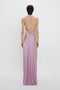 Rear view of a woman standing against a white background, wearing a long, elegant pastel purple camisole slip dress with thin straps and a crisscross back design from Victoria Beckham's Low Back Cami Floor-Length Dress In Rosa.