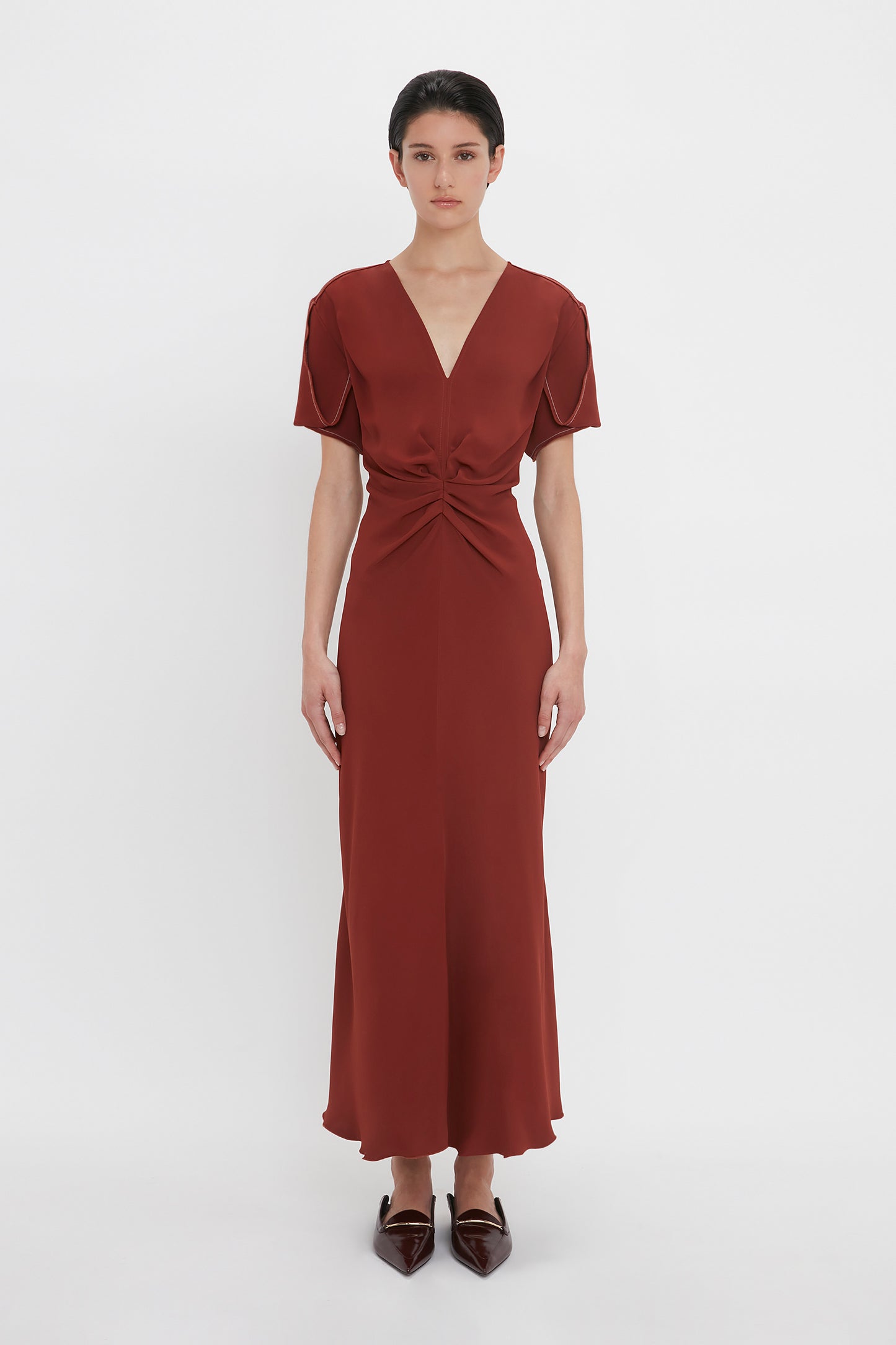 A person stands against a white background wearing a Gathered V-Neck Midi Dress In Russet by Victoria Beckham. The short-sleeved dress, made from figure-flattering stretch fabric, features a waist-defining pleat detail and knotted accent. The individual has short, dark hair and pointed shoes.
