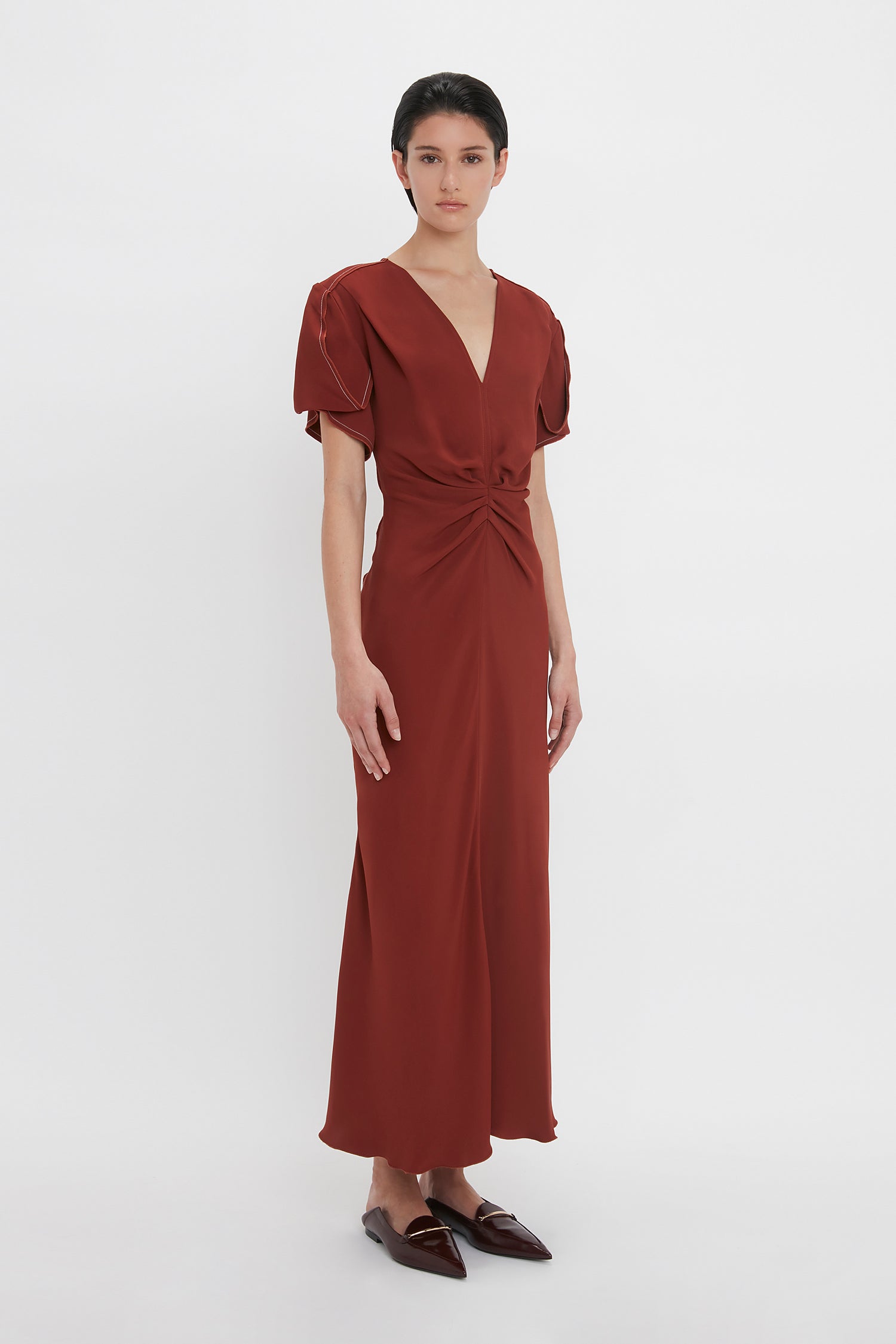 A person stands against a plain background, wearing a rust-colored Gathered V-Neck Midi Dress In Russet by Victoria Beckham with a waist-defining pleat detail and knotted at the waist, paired with dark brown flat shoes.