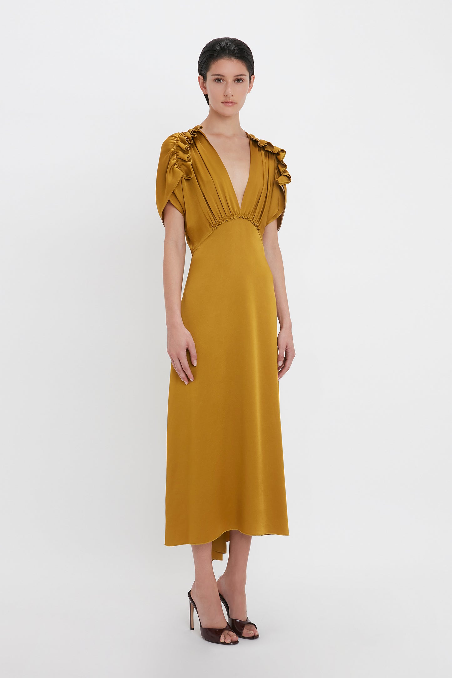 A person wearing a Victoria Beckham V-Neck Ruffle Midi Dress In Harvest Gold with short ruffled sleeves and black high-heeled shoes stands against a plain white background, exuding a seductive quality.