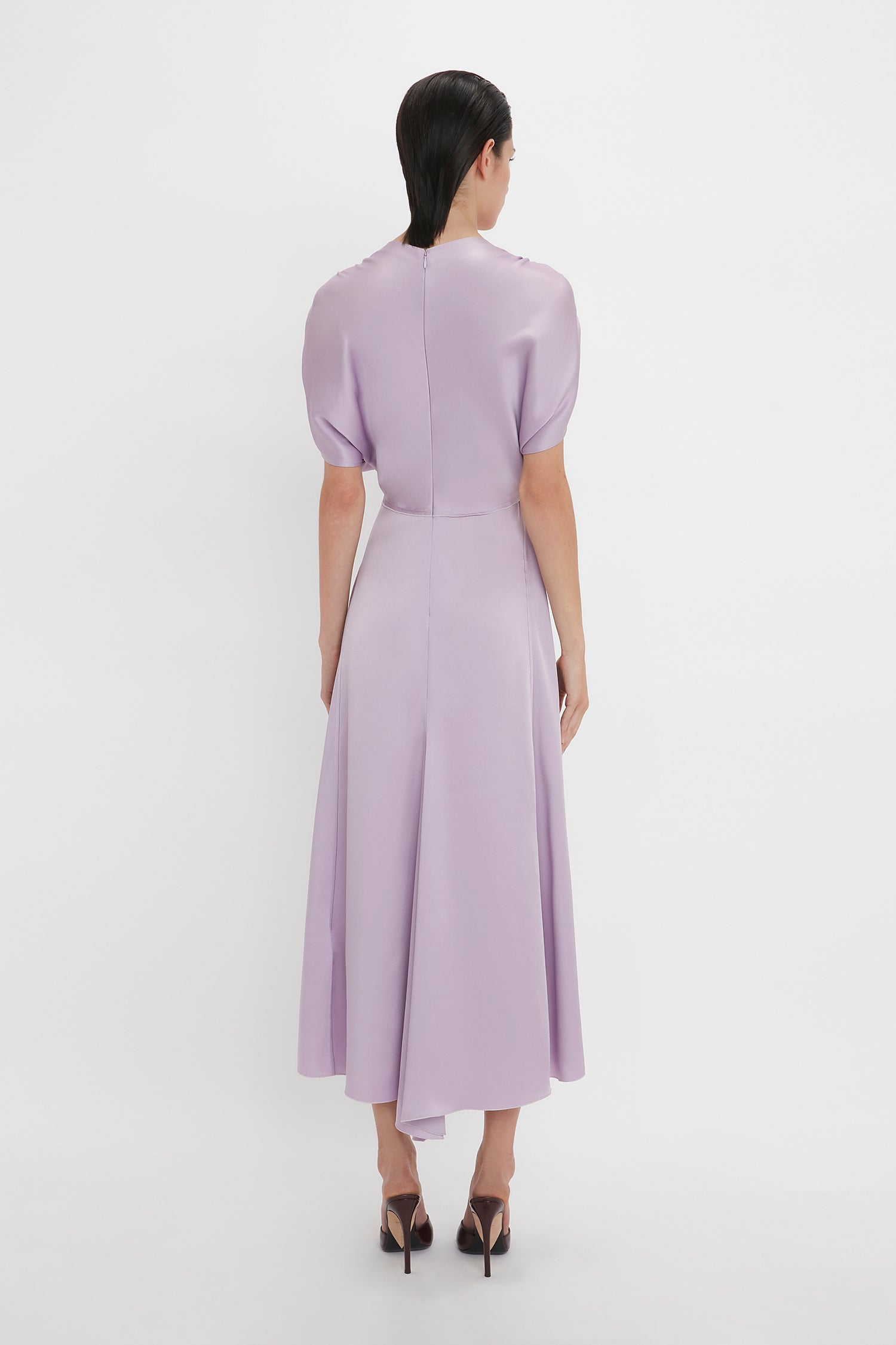 A person is standing facing away from the camera, wearing a Victoria Beckham V-Neck Ruffle Midi Dress In Petunia and black high-heeled shoes. The background is plain white, adding to the elegance reminiscent of Victoria Beckham's timeless style.