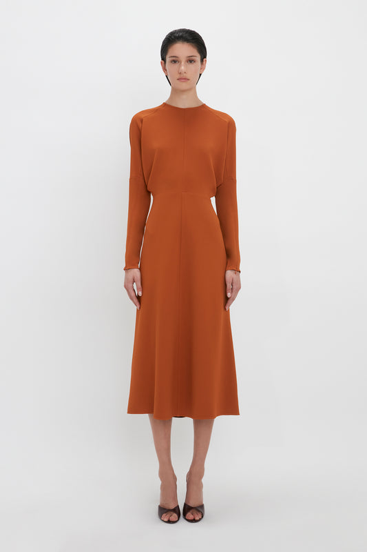 A woman in a plain, Victoria Beckham Dolman Midi Dress In Russet stands facing the camera, against a white background. She wears black open-toe heels.