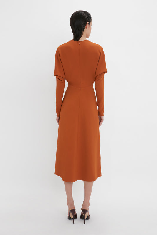 A woman stands facing away from the camera, wearing a Victoria Beckham Dolman Midi Dress In Russet with long sleeves and a high collar, paired with black high heels on a white background.