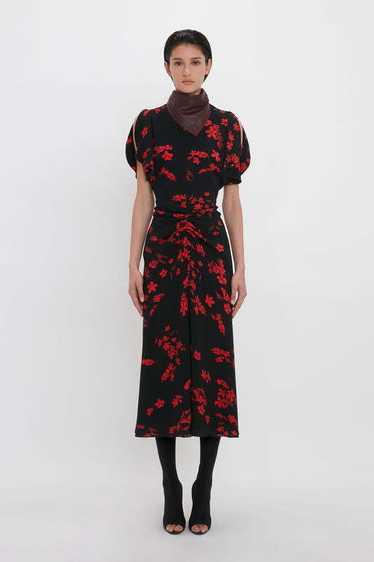 A person wearing a Gathered Waist Midi Dress In Sci-Fi Black Floral by Victoria Beckham, black open-toe shoes, and a dark brown scarf stands against a plain white background.