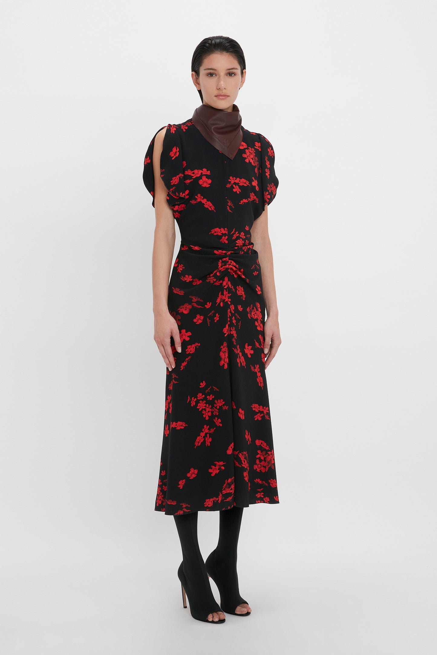A person stands wearing a Victoria Beckham Gathered Waist Midi Dress In Sci-Fi Black Floral, black tights, black open-toe heels, and a dark scarf around their neck. Their hands are by their sides against a plain white background.