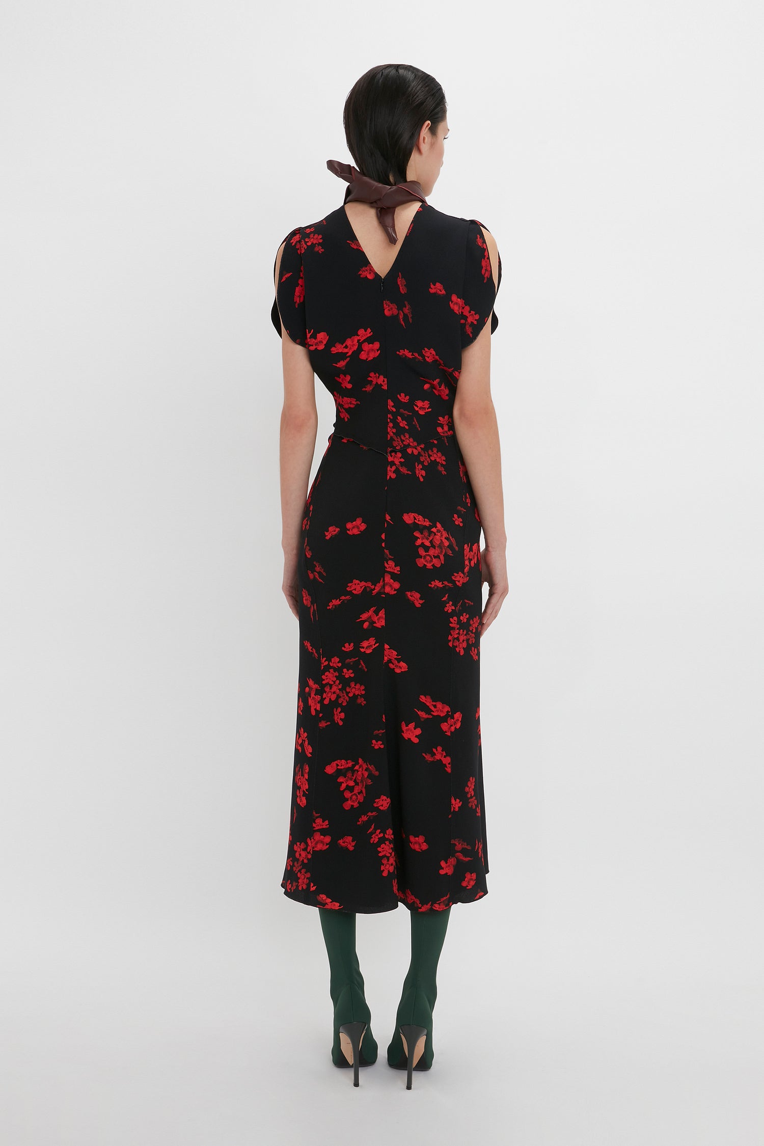 A person stands with their back facing the camera, wearing a black dress with red floral patterns and a fit-and-flare silhouette, complemented by green tights and black high-heeled shoes. The Gathered Waist Midi Dress In Sci-Fi Black Floral by Victoria Beckham features a keyhole back detail and is finished with a brown bow tie.